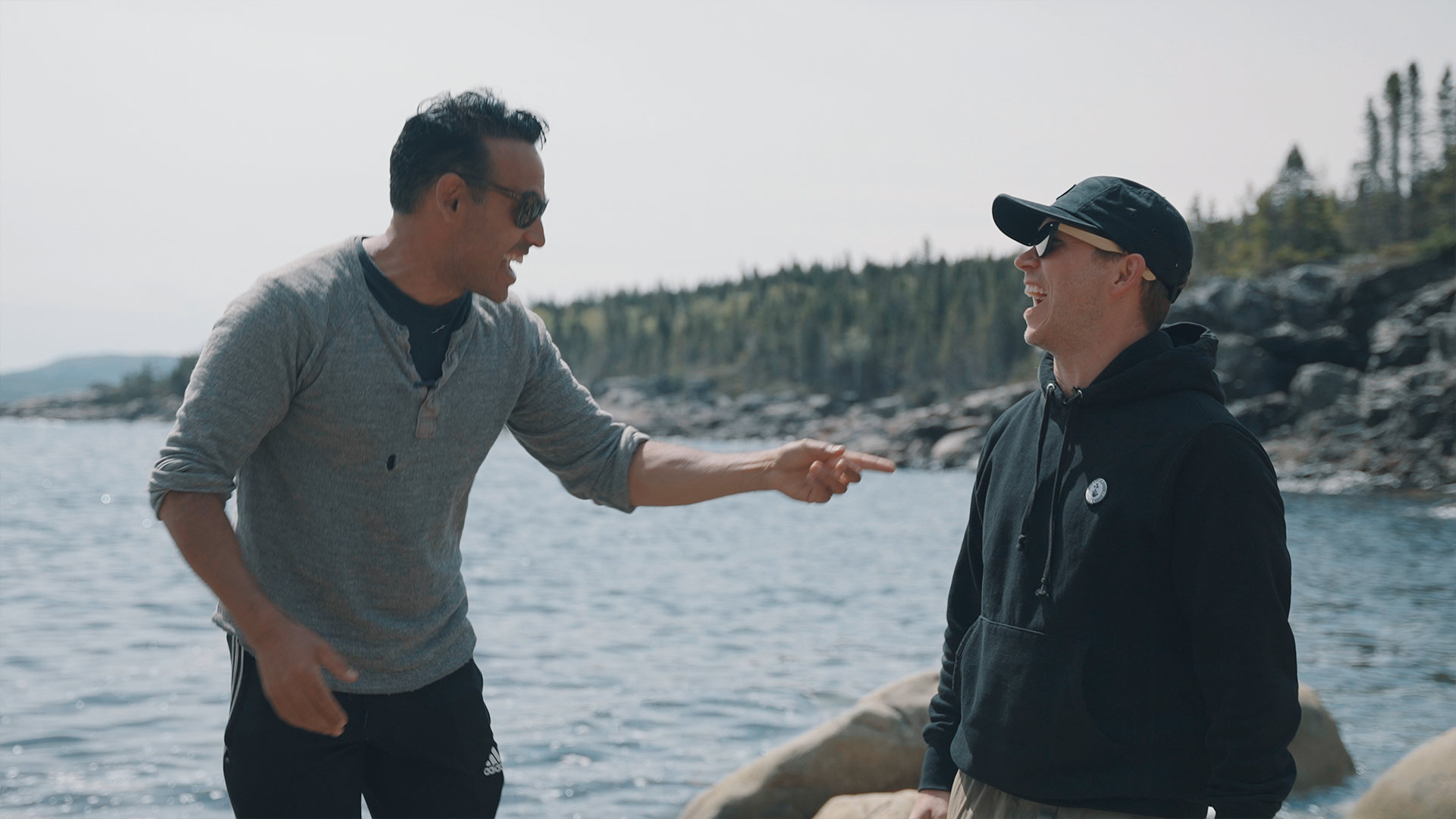 Shaun Majumder, left, and Dr. Michael Wahl, right, look at each other and laugh. There are water and trees in the image background.